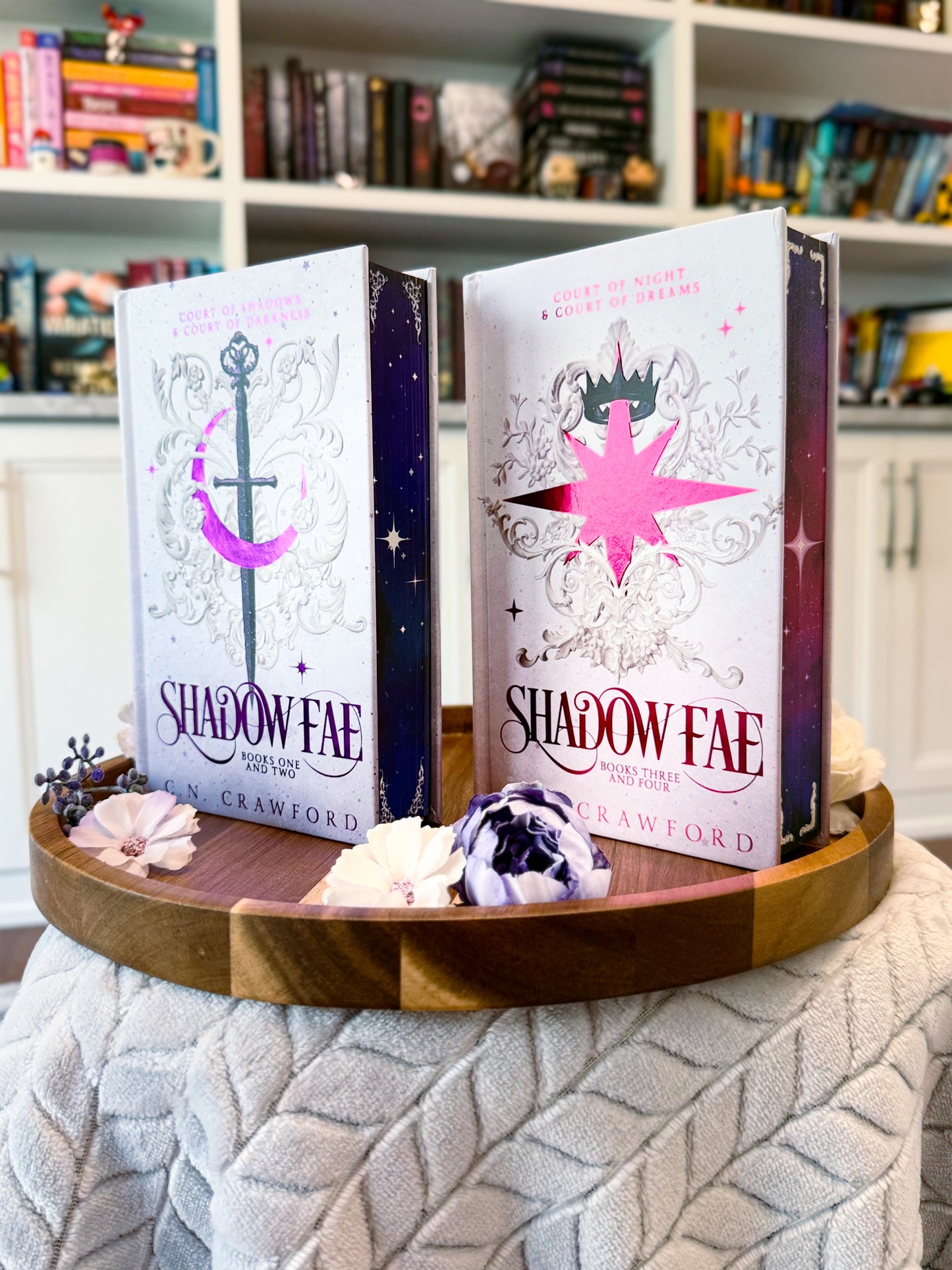 Two fancy special editions novels titled 'Shadow Fae 1-2' and 'Shadow Fae 3-4'  by C.N. Crawford. Books are displayed on a wooden tray with decorative items, against a bookshelf background.