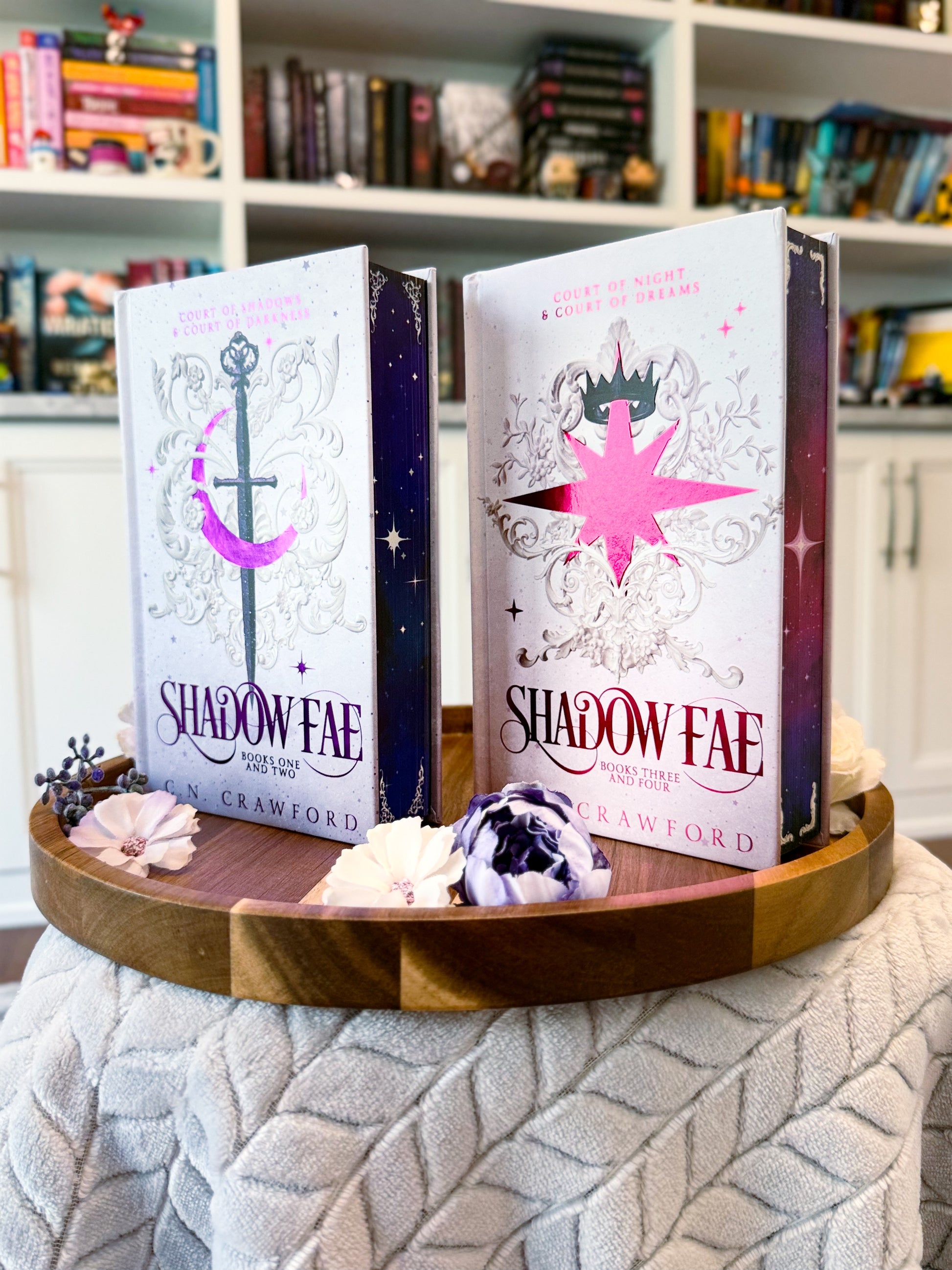 Two fancy special editions novels titled 'Shadow Fae 1-2' and 'Shadow Fae 3-4'  by C.N. Crawford. Books are displayed on a wooden tray with decorative items, against a bookshelf background.